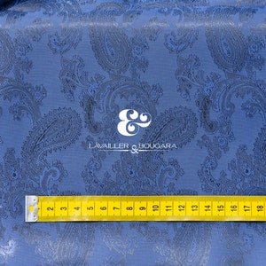 May include: Blue paisley patterned fabric with a yellow measuring tape across the bottom. The tape reads from 2 to 23 inches. The fabric has a subtle sheen and a textured surface.