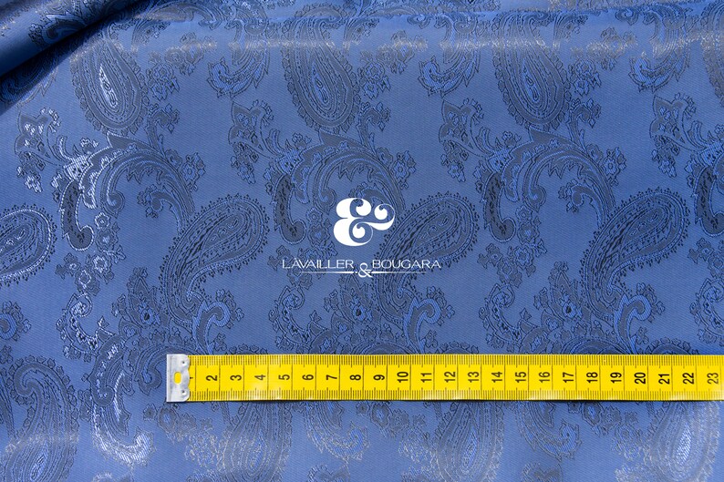 May include: Blue paisley patterned fabric with a yellow measuring tape across the bottom. The tape reads from 2 to 23 inches. The fabric has a subtle sheen and a textured surface.