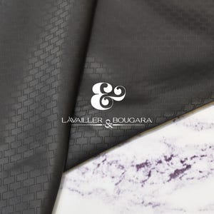 Może przedstawiać: Ciemnoszara tkanina z subtelnym geometrycznym wzorem, złożona, aby odsłonić kontrastującą białą i fioletową powierzchnię marmuru. Logo "LAVAILLER & BOUGARA" jest wyświetlane na biało. Tekstura i kolor tkaniny sugerują wysokiej jakości materiał.