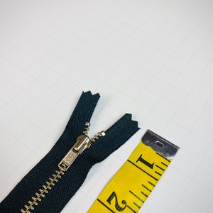May include: A black zipper with a silver slider, measured against a yellow tape measure. The tape measure shows the zipper is 1 inch long.