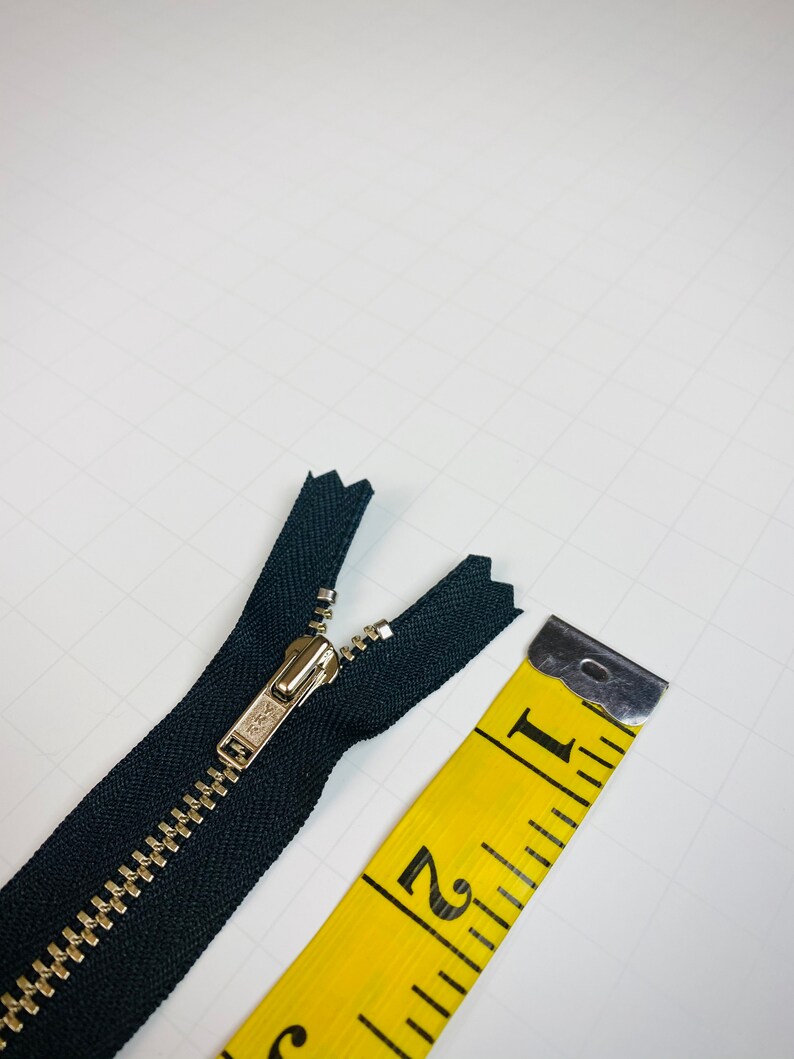 May include: A black zipper with a silver slider, measured against a yellow tape measure. The tape measure shows the zipper is 1 inch long.