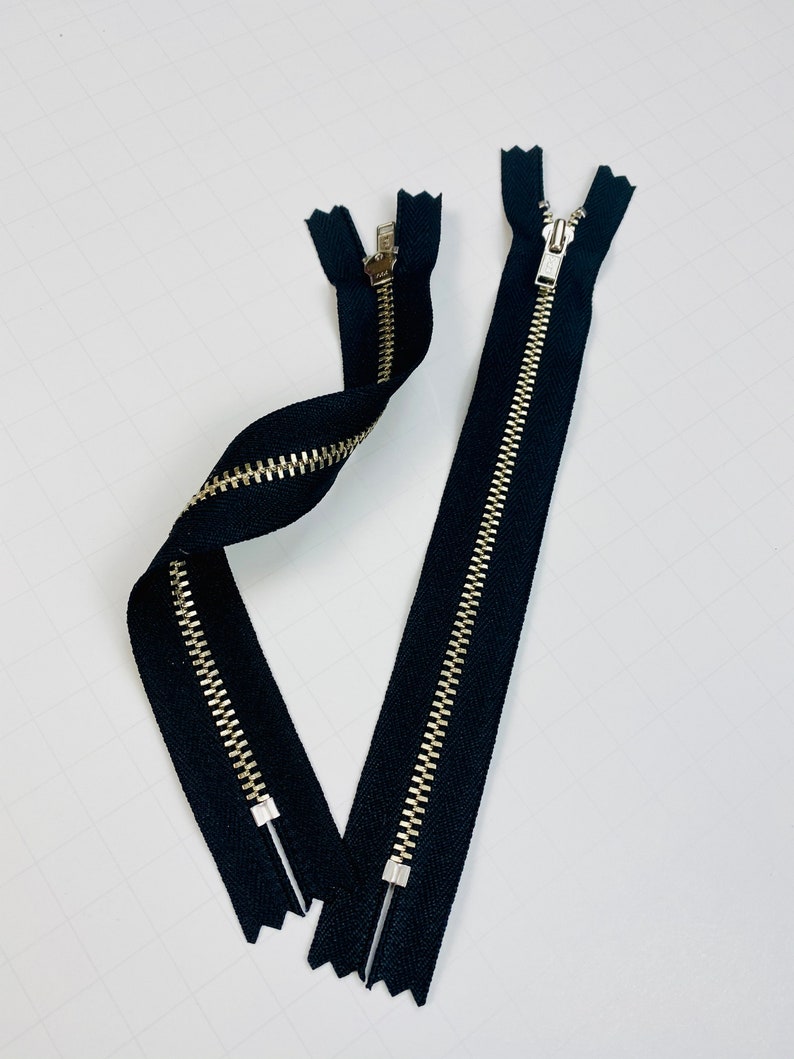 May include: Two black zipper closures with gold metal teeth. The zippers are laid out on a white surface.