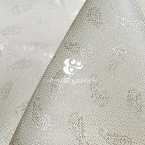 May include: Close-up of off-white fabric with a subtle paisley pattern. The fabric is folded, revealing the texture and design. The paisley motifs are in a lighter, metallic tone, creating a delicate contrast.