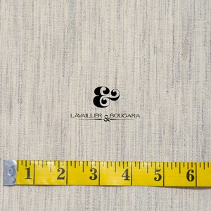 May include: A close-up of a beige fabric with a subtle texture. A yellow measuring tape with black markings is placed on the fabric, showing measurements from 1 to 8 inches. The fabric has a small black logo that reads "LAVAILLE & BOUGARDA".