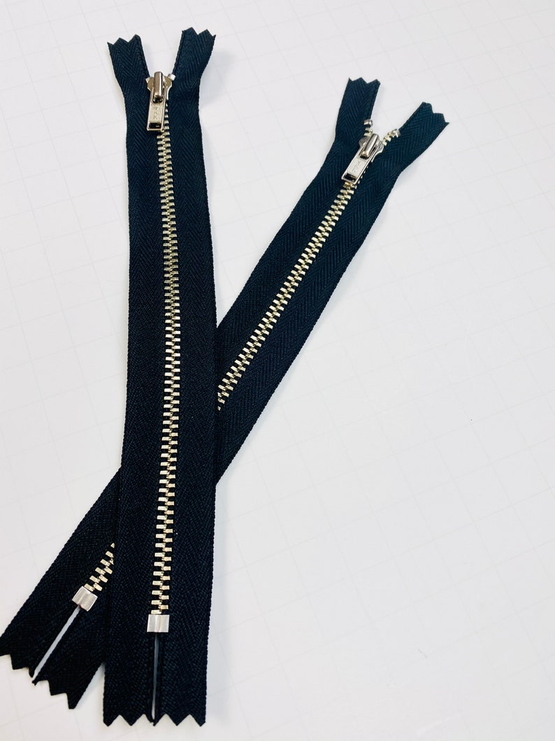 May include: Two black zipper closures with silver metal teeth. The zippers are laid out in a criss-cross pattern.