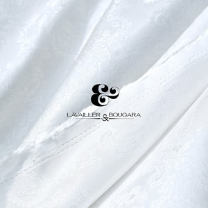 Może przedstawiać: Biała tkanina z wzorem paisley, z czarnym tekstem "LAVAILLER & BOUGARA". Tkanina jest udrapowana, ukazując fałdy i cienie, tworząc teksturowany wygląd. Ogólna estetyka jest elegancka i luksusowa.