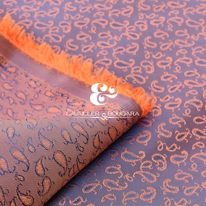 Puede incluir: Tela con estampado de cachemir en tonos morados y naranjas. La tela tiene una textura suave y un borde con flecos. El diseño de cachemir se repite en toda la superficie. El texto "LAVAILLER & BOUGARA" es visible.