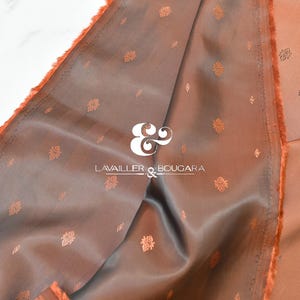 May include: Close-up of a folded fabric with a satin finish. The fabric is a muted orange color with a darker brown interior. Small, repeating floral patterns are visible. The brand name "LAVAILLER & BOUGARA" is printed in white.