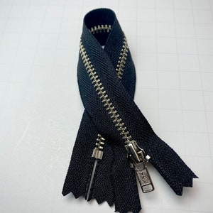 May include: A black zipper with a silver metal slider and teeth. The zipper is folded in half and the slider is at the top.