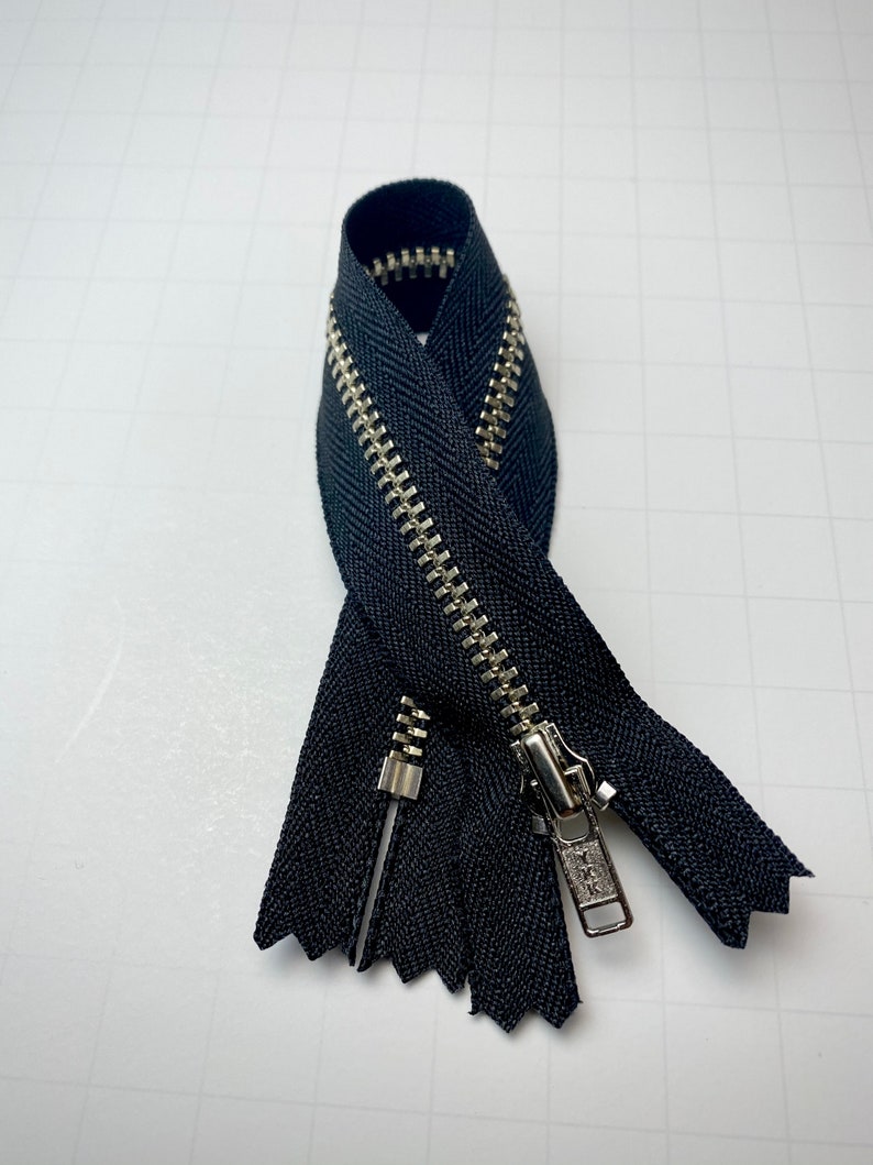 May include: A black zipper with a silver metal slider and teeth. The zipper is folded in half and the slider is at the top.