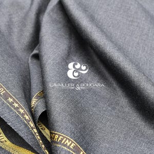 Puede incluir: Primer plano de tela gris con apariencia texturizada. Se ve una banda dorada con el texto "AUSTRALIAN MERINO WOOL" y "SUPERFINE". La tela está drapeada y el logotipo "LAVAILLER & BOUGARA" está presente.