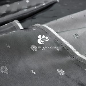 May include: Close-up of folded gray fabric with a subtle pattern and a textured appearance. The fabric has a white border and the text "LAURIER & BOUGARA" is visible. The fabric appears to be a high-quality textile.
