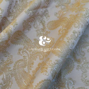 May include: Cream-colored fabric with a gold paisley pattern. The fabric is folded, showing the texture and design. The text "LAVAILLER & BOUGARA" is printed on the fabric.