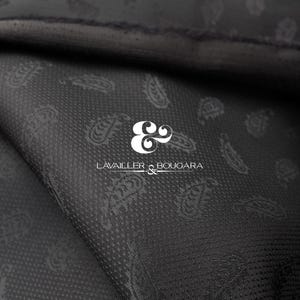 Può includere: Primo piano di un tessuto grigio scuro con un motivo paisley. Il tessuto ha una trama sottile ed è piegato, rivelando l'interno. Il testo bianco "LAVAILLER & BOUGARA" è visibile.