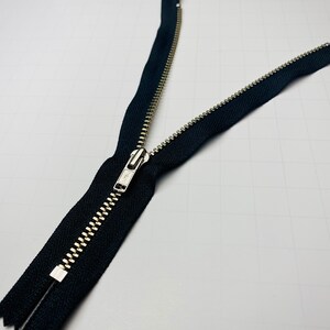 May include: A black zipper with a silver metal slider. The zipper is open and the two sides are separated.