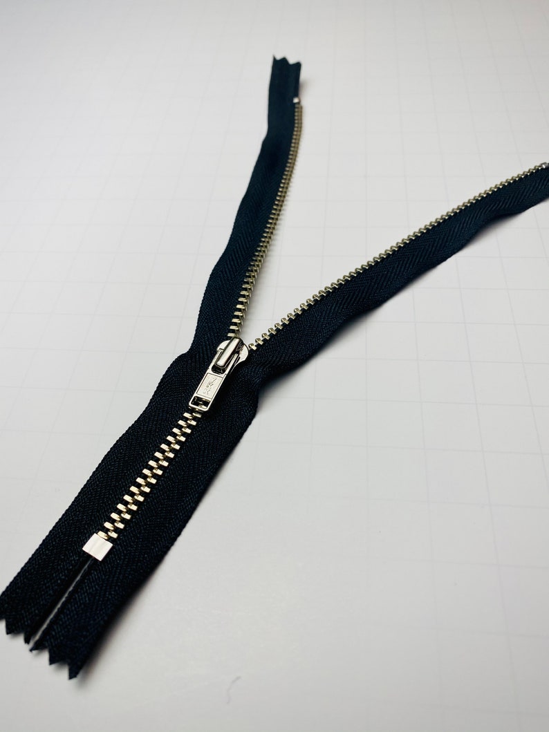 May include: A black zipper with a silver metal slider. The zipper is open and the two sides are separated.