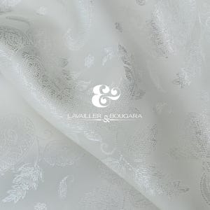 May include: White fabric with a subtle paisley pattern and a soft sheen. The fabric is draped, creating folds and shadows that highlight the design. The logo "Lavailler & Bougara" is visible.
