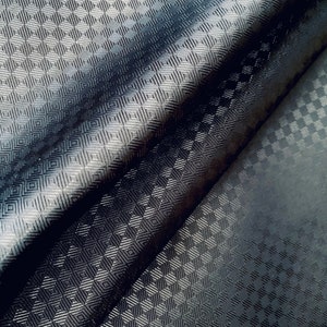 Puede incluir: Primer plano de una tela oscura con un patrón geométrico de diamantes repetido. La tela está doblada, creando sombras y luces que enfatizan la textura y la profundidad del diseño. La apariencia general es elegante y moderna.