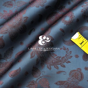 Puede incluir: Primer plano de una tela azul con un estampado de cachemira en tonos burdeos. Una cinta métrica amarilla está sobre la tela. La tela tiene el texto "LAVAILLER & BOUGARA" en blanco.