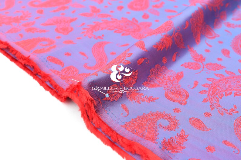 Puede incluir: Primer plano de una tela con un estampado de cachemira en tonos morados y coral. La tela tiene un borde sin rematar y deshilachado en coral. El texto "LAVAILLER & BOUGARA" es visible en la tela.