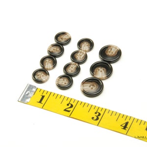 May include: A set of ten brown and beige plastic buttons with a swirl pattern. The buttons are arranged in two rows of five. A yellow measuring tape with black numbers is in the foreground.