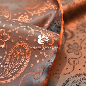 Puede incluir: Primer plano de una tela con un estampado de cachemira en tonos marrón y naranja. La tela tiene un brillo satinado y un diseño floral detallado. El logotipo "AVALLER & BOUCARA" es visible.