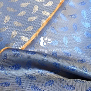 May include: Close-up of blue fabric with a paisley pattern in silver and blue. The fabric has a textured appearance and a gold-colored trim. The logo "LAVALLER & BOUGARA" is visible.