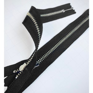 May include: Two black zipper tapes with silver metal teeth. The zippers are laid out on a white surface.