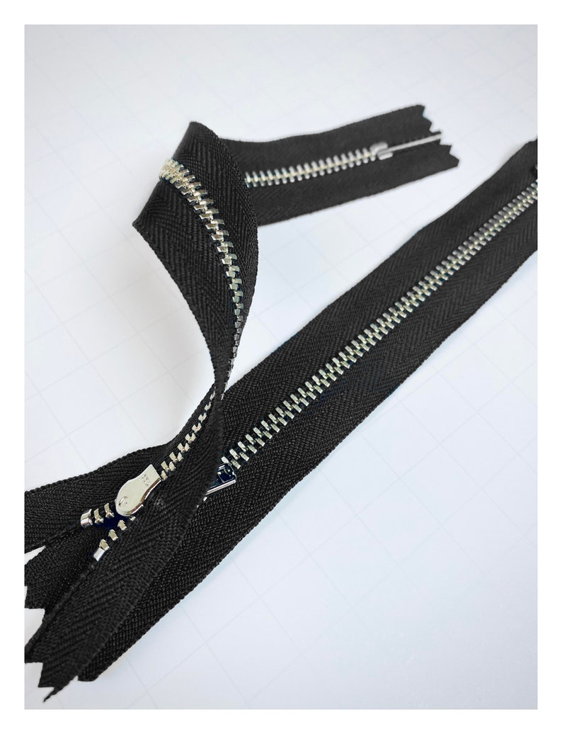 May include: Two black zipper tapes with silver metal teeth. The zippers are laid out on a white surface.