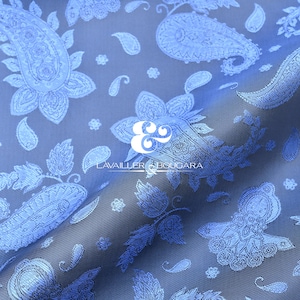 May include: A close-up of a blue fabric with a paisley pattern. The design features large, detailed paisley shapes, smaller teardrop motifs, and floral elements. The fabric has a subtle sheen, and the overall effect is elegant and decorative.