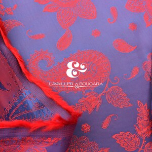 Puede incluir: Primer plano de un textil con un estampado de cachemira en tonos rojos y azules. La tela tiene un aspecto texturizado con el texto "LAVAILLER & BOUGARA" en blanco. Una franja roja es visible a lo largo del borde de la tela.