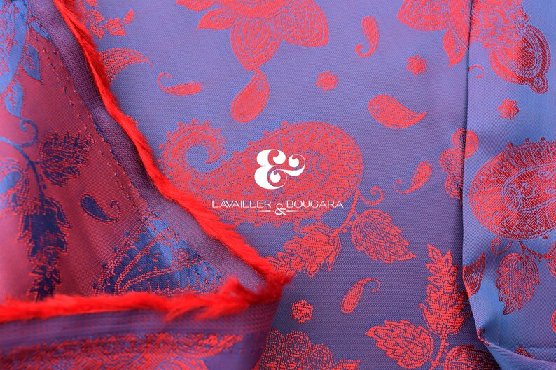 Puede incluir: Primer plano de un textil con un estampado de cachemira en tonos rojos y azules. La tela tiene un aspecto texturizado con el texto "LAVAILLER & BOUGARA" en blanco. Una franja roja es visible a lo largo del borde de la tela.