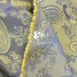 May include: A close-up of a fabric with a paisley pattern in shades of blue, gold, and brown. The fabric has a textured surface and a fringed edge.