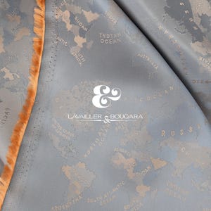 May include: Close-up of a folded fabric with a world map pattern in beige on a light blue background. The edge of the fabric has a fringed trim in a warm orange hue. The brand name "LAVAILLER & BOUGARA" is visible.