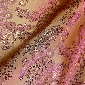 Könnte beinhalten: Nahaufnahme eines Stoffes mit Paisley-Muster. Der Stoff ist hellbraun mit einem pinkfarbenen und goldenen Metallic-Garn, das in das Design eingewebt ist.