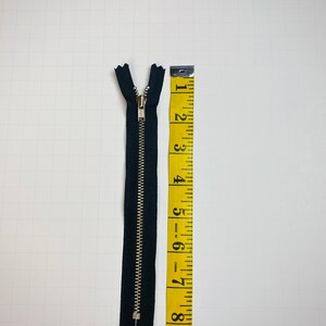 May include: A black zipper with a silver slider, measuring 6 inches long, next to a yellow measuring tape.