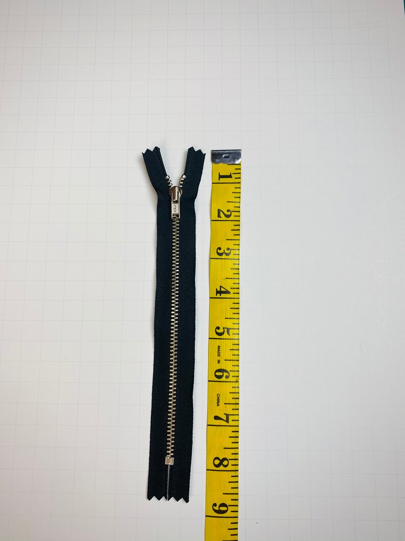 May include: A black zipper with a silver slider, measuring 6 inches long, next to a yellow measuring tape.