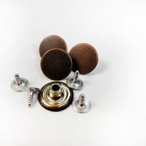 May include: A collection of bronze-colored metal jean buttons and silver-colored screws. The buttons are round with a slightly textured surface. The screws have a slotted head. The items are arranged on a white surface.