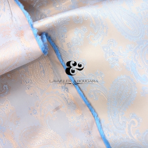 May include: Close-up of a light pink fabric with a blue paisley pattern. The fabric is folded over and the edges are trimmed with a blue fringe. The text "LAVAILLEUR & BOUGARA" is visible in the center of the image.