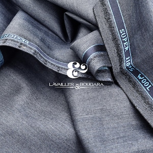 Puede incluir: Primer plano de una tela de lana gris oscuro doblada, con un sutil patrón de espiga. Una etiqueta a rayas azules y blancas dice "SUPER 110'S WOOL EXCELLENCE". La tela es de la marca "LAVAILLER & BOUGARA".