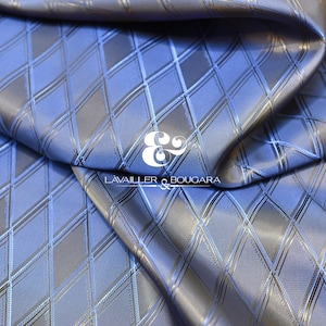 Puede incluir: Primer plano de una tela azul y gris con un patrón de diamantes. La tela tiene un brillo sutil y está doblada, creando sombras y reflejos. El texto "LAVAILLER & BOUGARA" es visible.