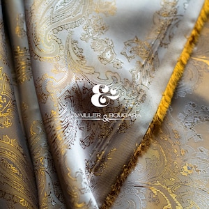 Puede incluir: Primer plano de una tela blanca y dorada con un patrón de paisley. La tela tiene una franja dorada a lo largo del borde. La tela está etiquetada "E & E, VAILLER & BOUGARE"