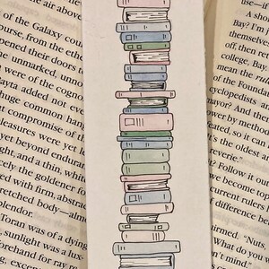 May include: A hand-drawn illustration of a stack of books in various colours, including pink, blue, and green. The books are stacked on top of each other, with the top book being the smallest and the bottom book being the largest. The illustration is done in a simple, whimsical style.