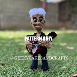 May include: Crochet pattern for a musician doll holding a red electric guitar. The doll has white hair, a brown face, and is wearing a black shirt and blue jeans. The pattern is available for purchase from @GEEKYCAKESANDCRAFTS.