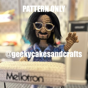 May include: Crochet pattern for a musician doll with long black hair, wearing sunglasses and a blue and white plaid shirt, standing in front of a white keyboard with the word "Mellotron" printed on it. The pattern is available on the website @geekycakesandcrafts.