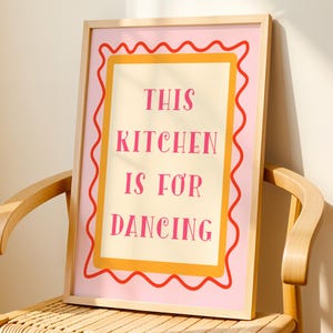 Może przedstawiać: Wydruk w różowej i żółtej ramie z tekstem "This kitchen is for dancing" w różowym i białym kolorze czcionki.