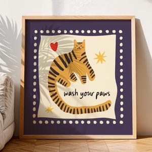 K&ouml;nnte beinhalten: Ein quadratisches gerahmtes Wandbild mit einer beigefarbenen Katze mit dunkelbraunen Streifen auf cremefarbenem Hintergrund. Die Katze ist C-f&ouml;rmig gebogen, darunter steht der Text "wash your paws". Ein rotes Herz und zwei gelbe Sterne umgeben die Katze, alles umschlossen von einem dunkelvioletten Rand mit wei&szlig;en Punkten.