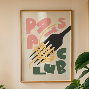 May include: A dining room scene featuring a wooden table and chairs, a large framed poster with the words "Pasta Club", and several potted plants. A black pendant light hangs above the table, and a wooden cabinet with plants sits in the background.