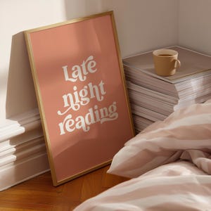 May include: A framed poster with the words "Late night reading" in white script on a peach background. A tan mug sits on a stack of papers, and a striped comforter is in the foreground.