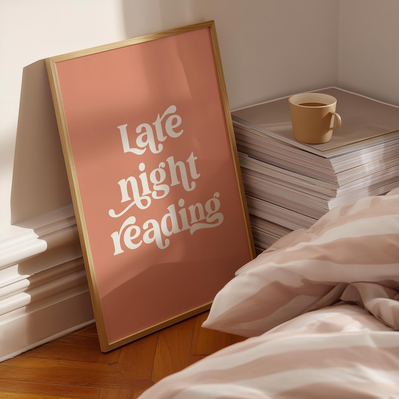 May include: A framed poster with the words "Late night reading" in white script on a peach background. A tan mug sits on a stack of papers, and a striped comforter is in the foreground.